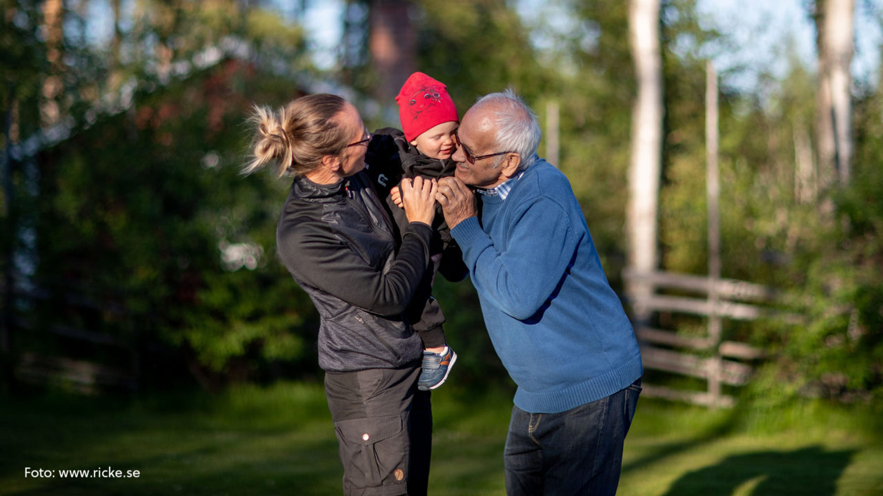Generationsbild. Litet barn ger en kram till morfar/farfar. Mamma håller i barnet.