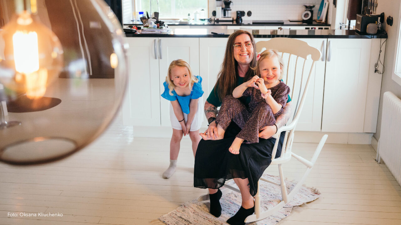 Mamma som håller i en dotter medan hon sitter i gungstolen och den andra står bredvid.