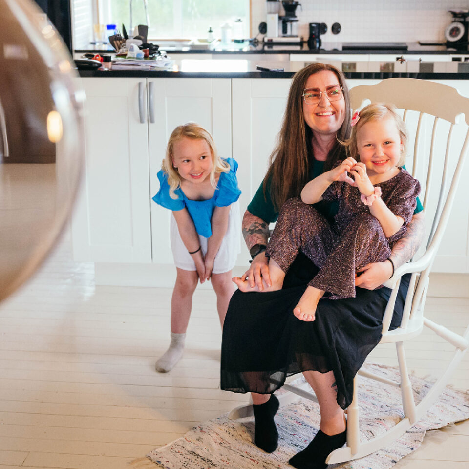 Mamma som håller i en dotter medan hon sitter i gungstolen och den andra står bredvid.